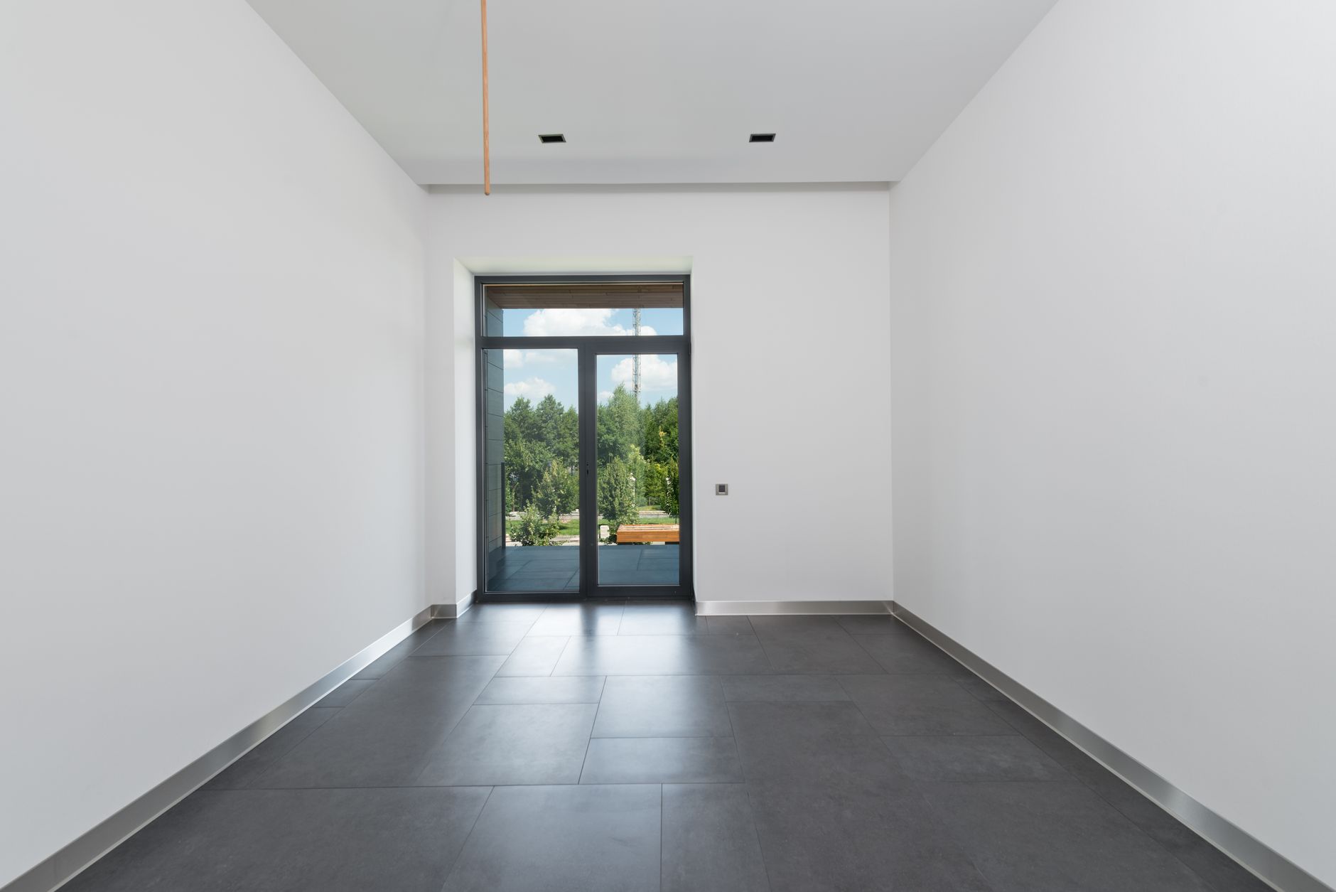 Salle vide aux murs blancs et au sol gris foncé, avec une porte vitrée donnant sur une terrasse et des arbres.
