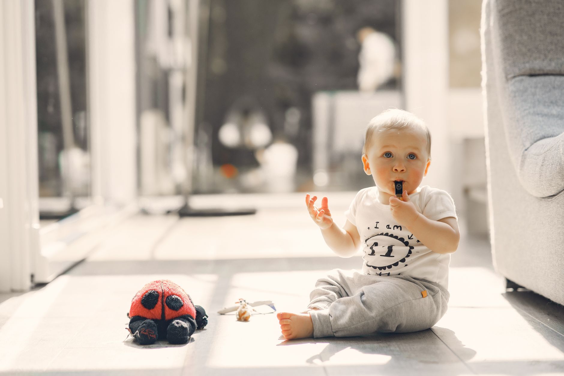 Un bébé assis sur un sol clair mord un petit objet noir, avec une peluche coccinelle rouge et noire posée devant lui.